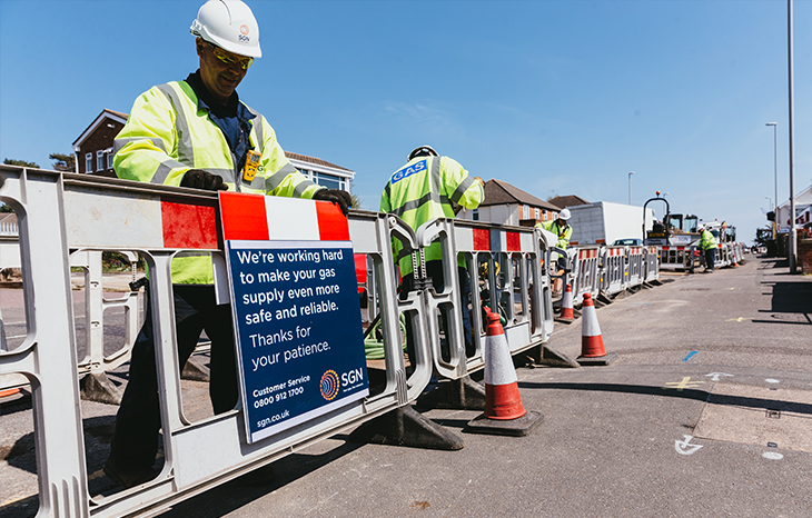 An engineer placing a sign on some roadworks barriers. The sign reads: We're working hard to make your gas supply even more safe and reliable. Thanks for your patience.