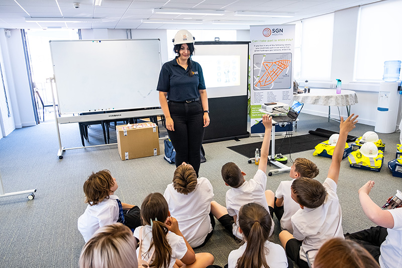 A woman wearing a hard hat demonstrating to school children sitting on the floor