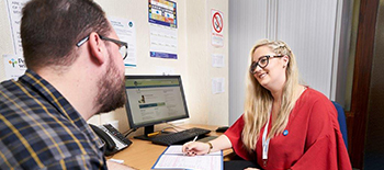 a woman sits at a desk with paperwork, she is talking to a man