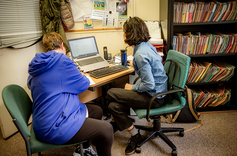 2 women sit at a desk, looking at a laptop screen