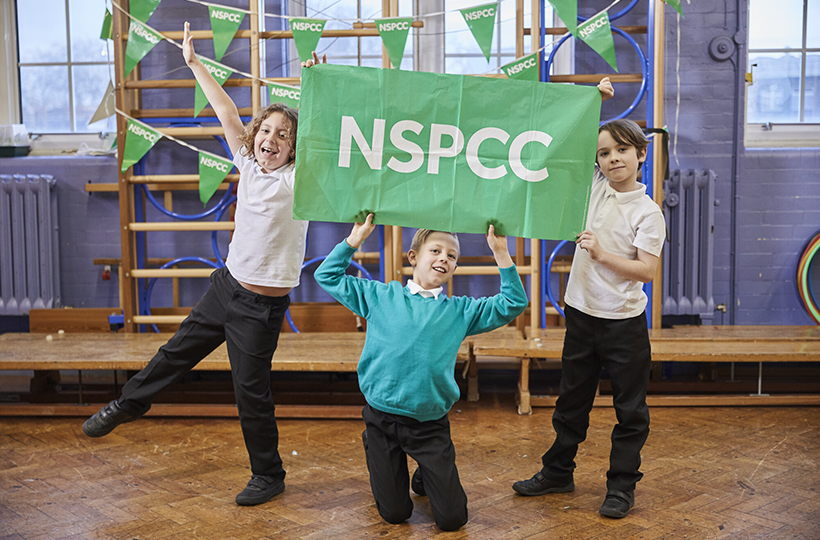 3 children in a school gym, holding up a large green sign reading NSPCC