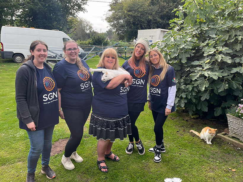 Five women in SGN t-shirts standing on grass, with one holding a white cat while a ginger cat stands next to them.