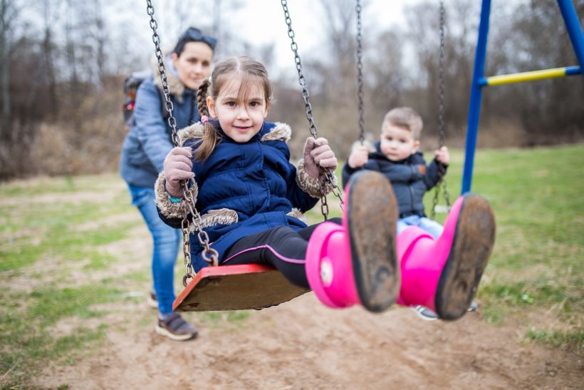 Two children are sitting on a swing with their parent in the background