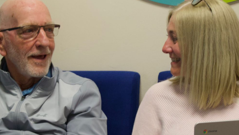 An older man is sitting and talking to a woman using a laptop