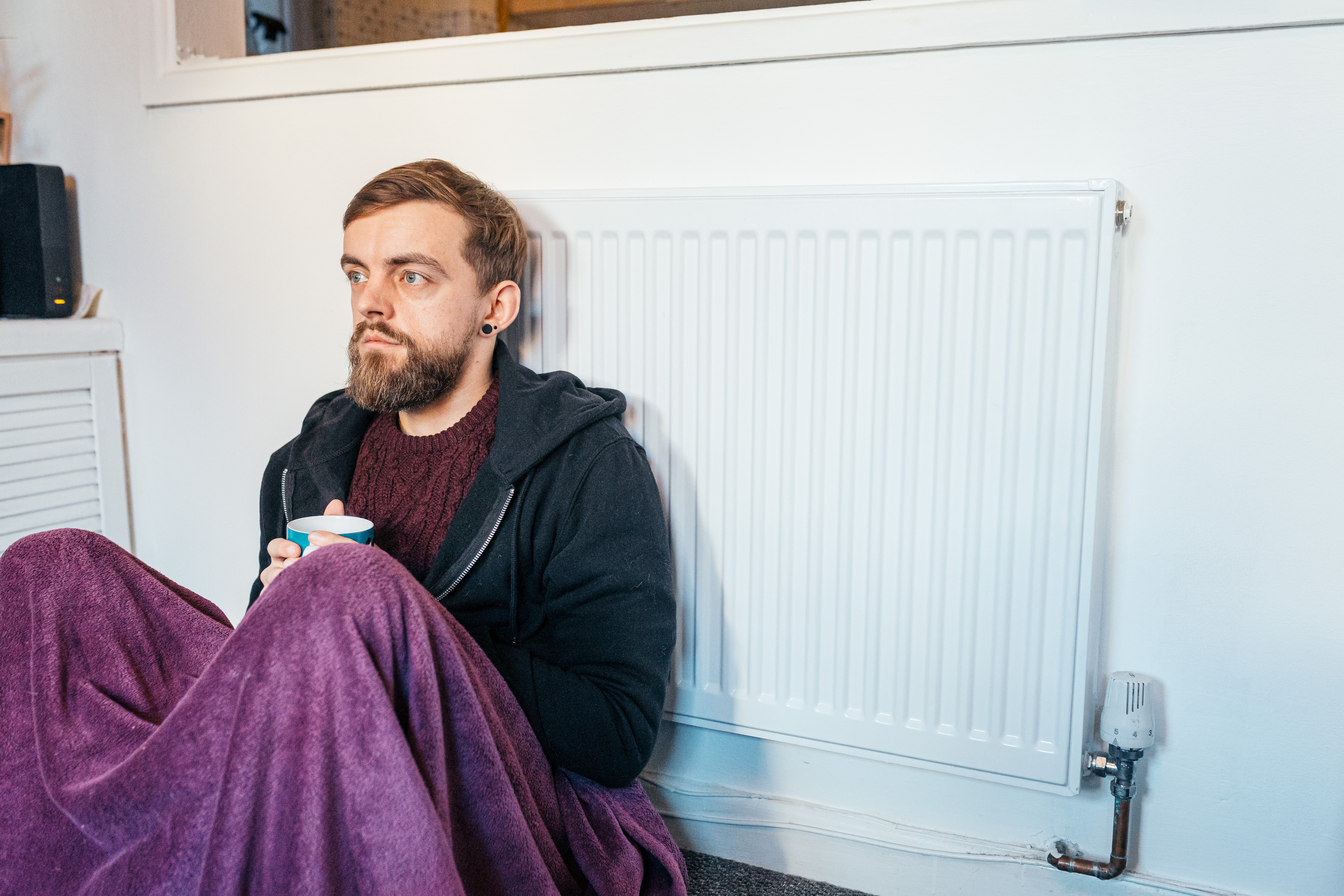 A young man covered by a blanket sat against a radiator