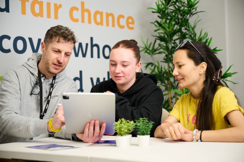 A YCMA adviser is sitting at a table in conversation with two young people. All three of them are looking at a tablet screen.. 