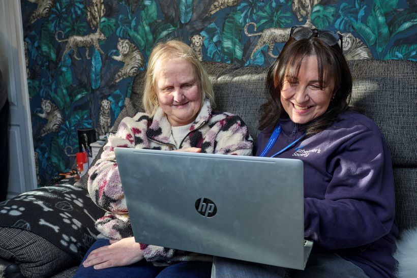 An Alzheimer's Society advisor is sitting on a sofa with an older lady, both looking at a laptop screen