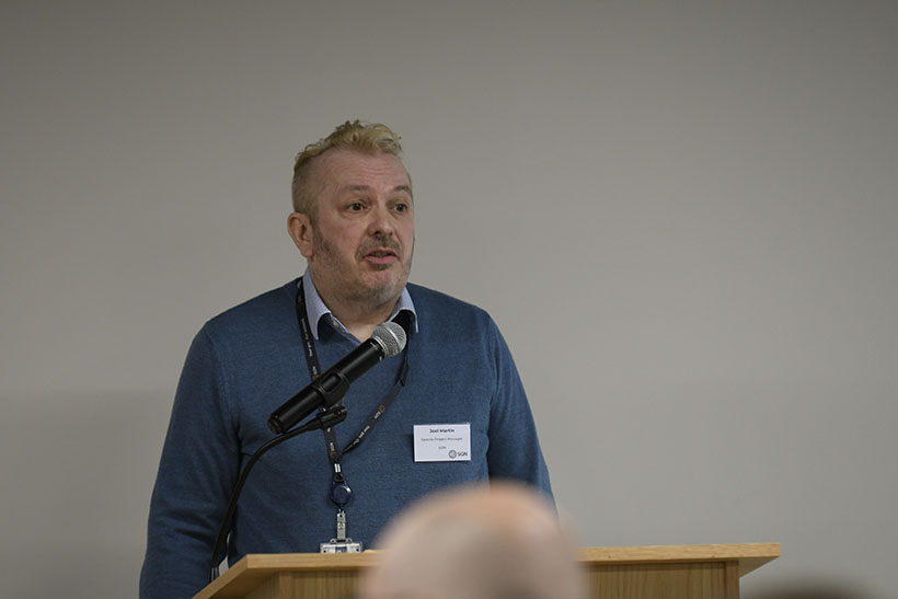 Head and shoulders image of man speaking at SGN event. He has a microphone in front of him as he addresses the audience.