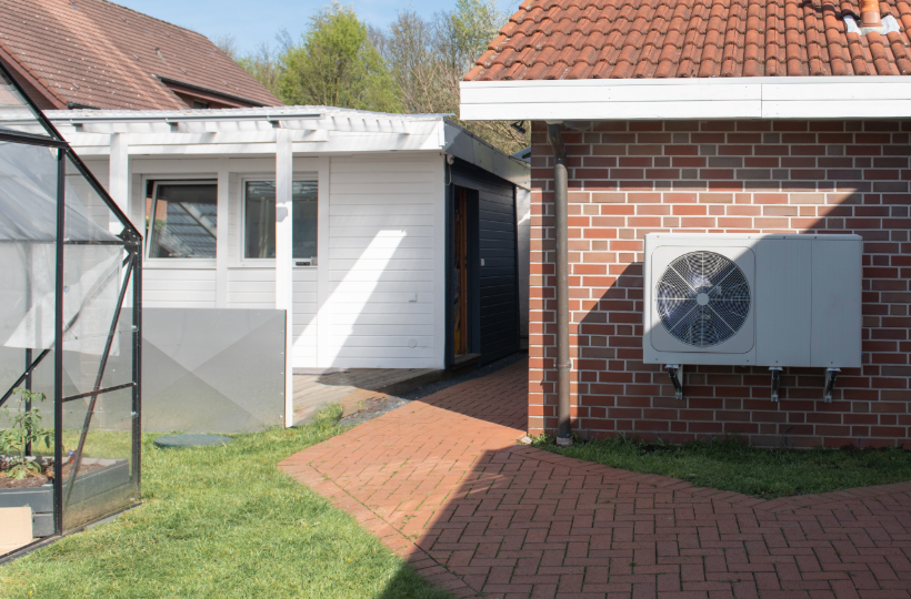 A heat pump installed on a garage wall. A greenhouse and grass can be seen next to the garage.