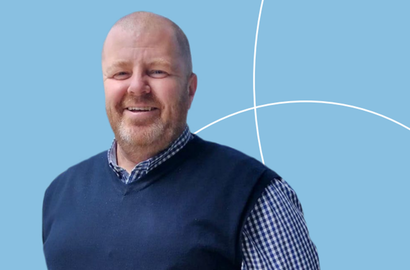 A man wearing a sleeveless blue jumper and a blue checked shirt. He is smiling.