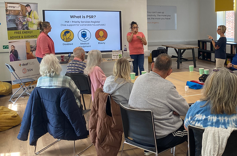 A community information session taking place in a bright meeting room. Several attendees are seated around tables facing a large presentation screen displaying information about the Priority Services Register (PSR), with icons for disability, medical equipment, and elderly support. Two presenters stand at the front of the room, one using sign language.