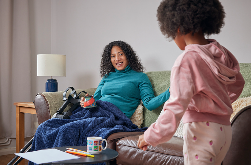 A person sitting on a sofa with a mobility aid nearby talks with a child standing in a living room at home.