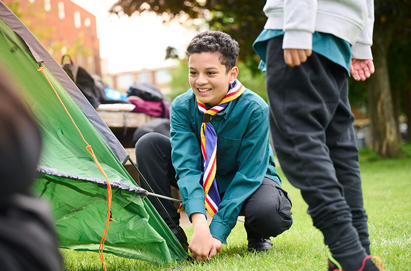 A young Scouts member pitches a green tent on a grassy field during an outdoor group activity.
