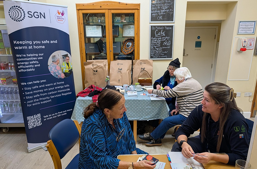 An adviser speaks with community members at a Warm Welcome Spaces hub, with an SGN banner displaying energy support and home safety information.
