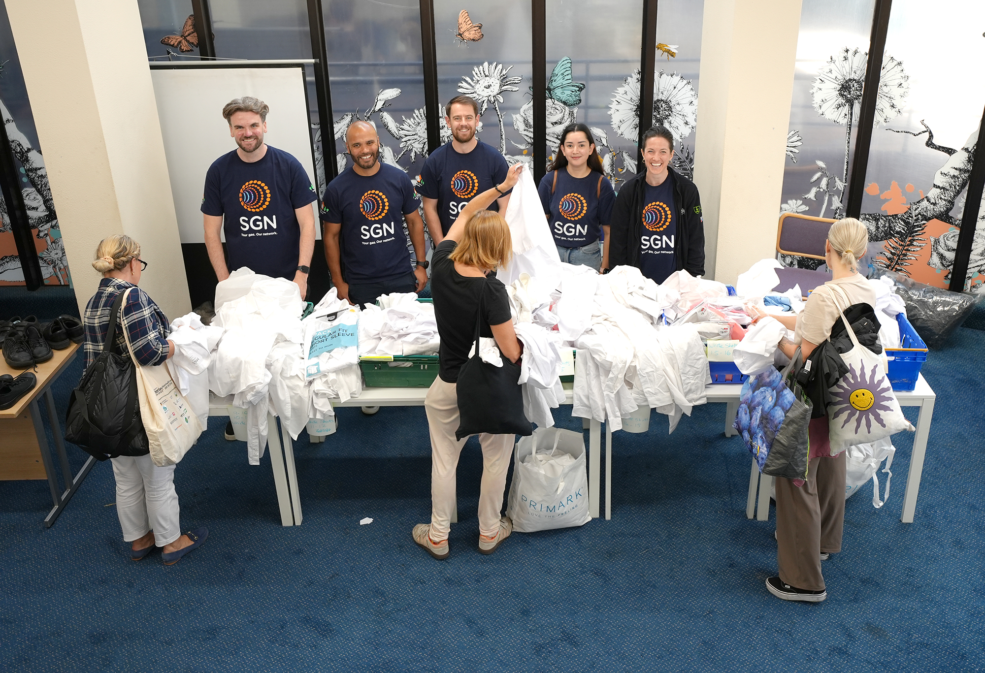 Five SGN volunteers smiling behind a table of school shirts as parents look through them