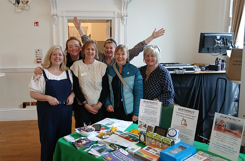 Six people stand behind an information stall at a community event in a large indoor hall. The table is covered with leaflets, brochures, energy-saving products and display materials. Several information boards on the table promote Alienergy services, including home energy efficiency and retrofit support. One person standing at the back has both arms raised.