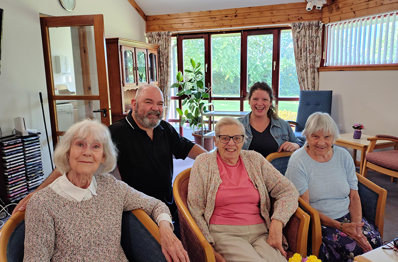Five people are seated and standing together in a bright communal lounge. Three older adults sit in armchairs at the front, while two other adults sit behind them. The room has large windows overlooking a garden, wooden ceiling panels, upholstered chairs, a display cabinet and potted plants, creating a comfortable indoor social space.