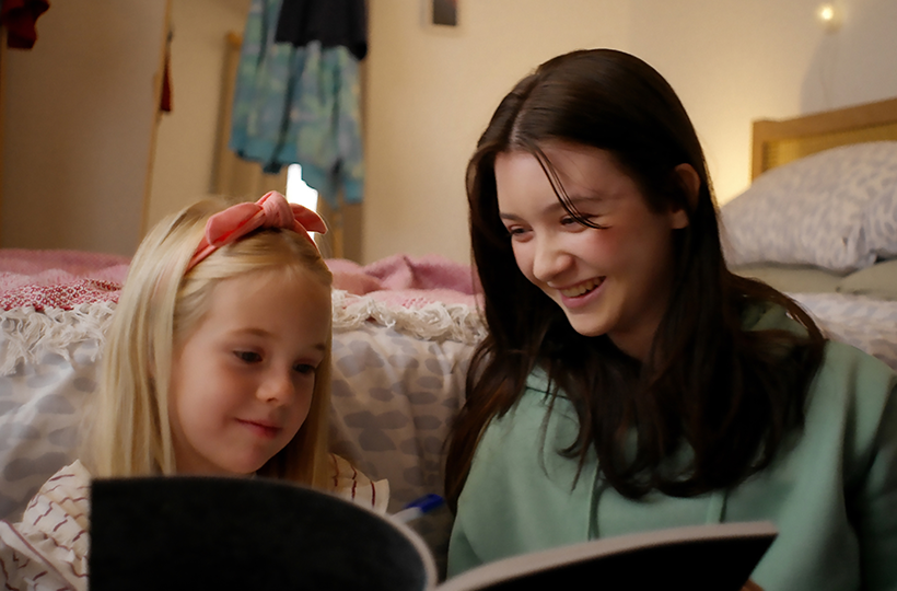 Woman with long dark hair and a young child with a pink headband sitting beside a bed, looking at and sharing a book together in a softly lit bedroom. Bedding and blankets are visible in the background.