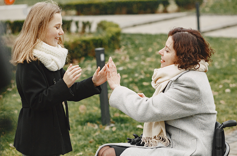 A child in a black coat and white knitted scarf stands facing a seated person wearing a light grey coat and cream scarf, who is using a wheelchair. Both have their hands raised and touching in a hand-clapping game or activity in a park setting. Grass, shrubs and pathways are visible in the background.