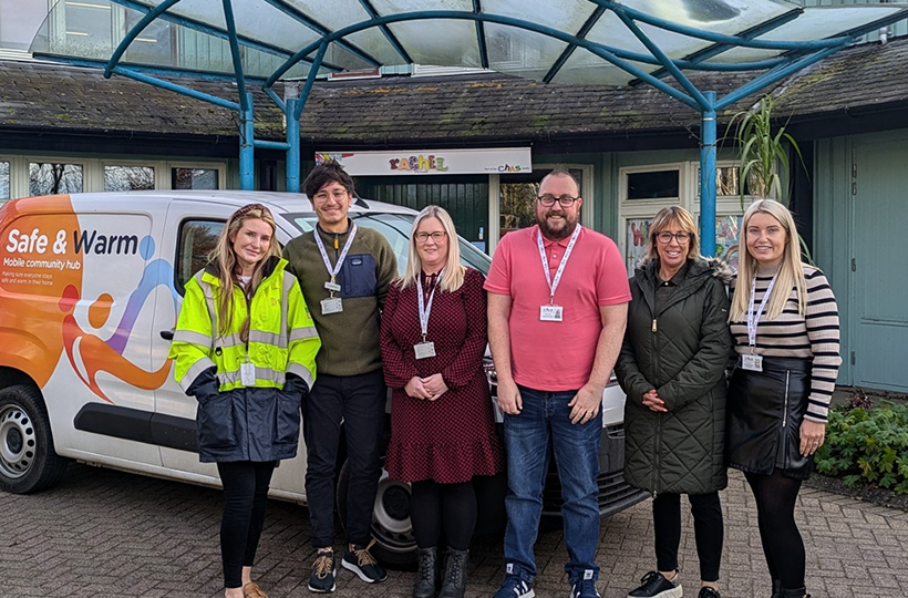 Six people stand together outside a community building in front of a white and orange “Safe & Warm” mobile community hub van. The group is gathered beneath a covered entrance and all are wearing staff ID lanyards. The van displays the text “Safe & Warm” and colourful graphics on its side.
