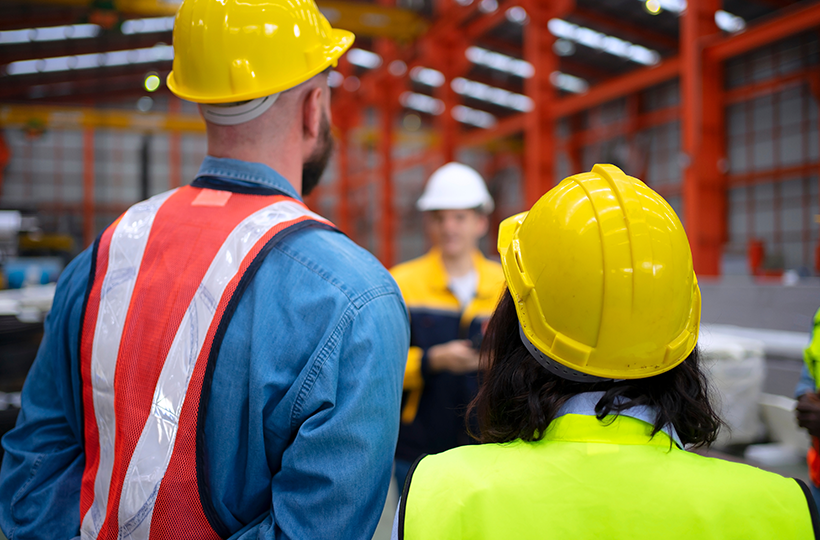 Two workers wearing yellow hard hats and high-visibility safety clothing stand in the foreground of an industrial facility, facing a small group during a workplace briefing. Red steel framework and factory equipment are visible in the background.