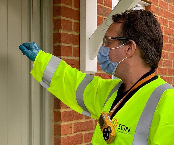 A SGN engineer wearing a high-viz jacket, a face mask and disposable gloves knocks on a customer's door.