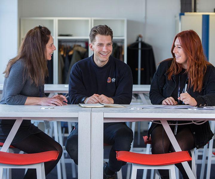 Three people in an office, sitting together at a desk. Everyone is smiling. 