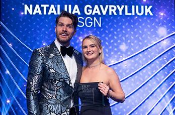 Tiera Box collects the Rising Star award on behalf of Nataliia Gavryliuk from host Joel Dummett. Nataliia's name is captioned on the screen behind the host in the background.