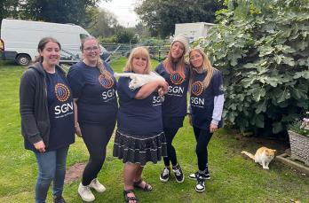 Five women in SGN t-shirts standing on grass, with one holding a white cat while a ginger cat stands next to them.