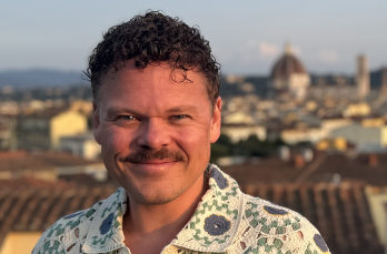 A smiling man with white skin, curly hair and moustache on the rooftop of a sunny European town