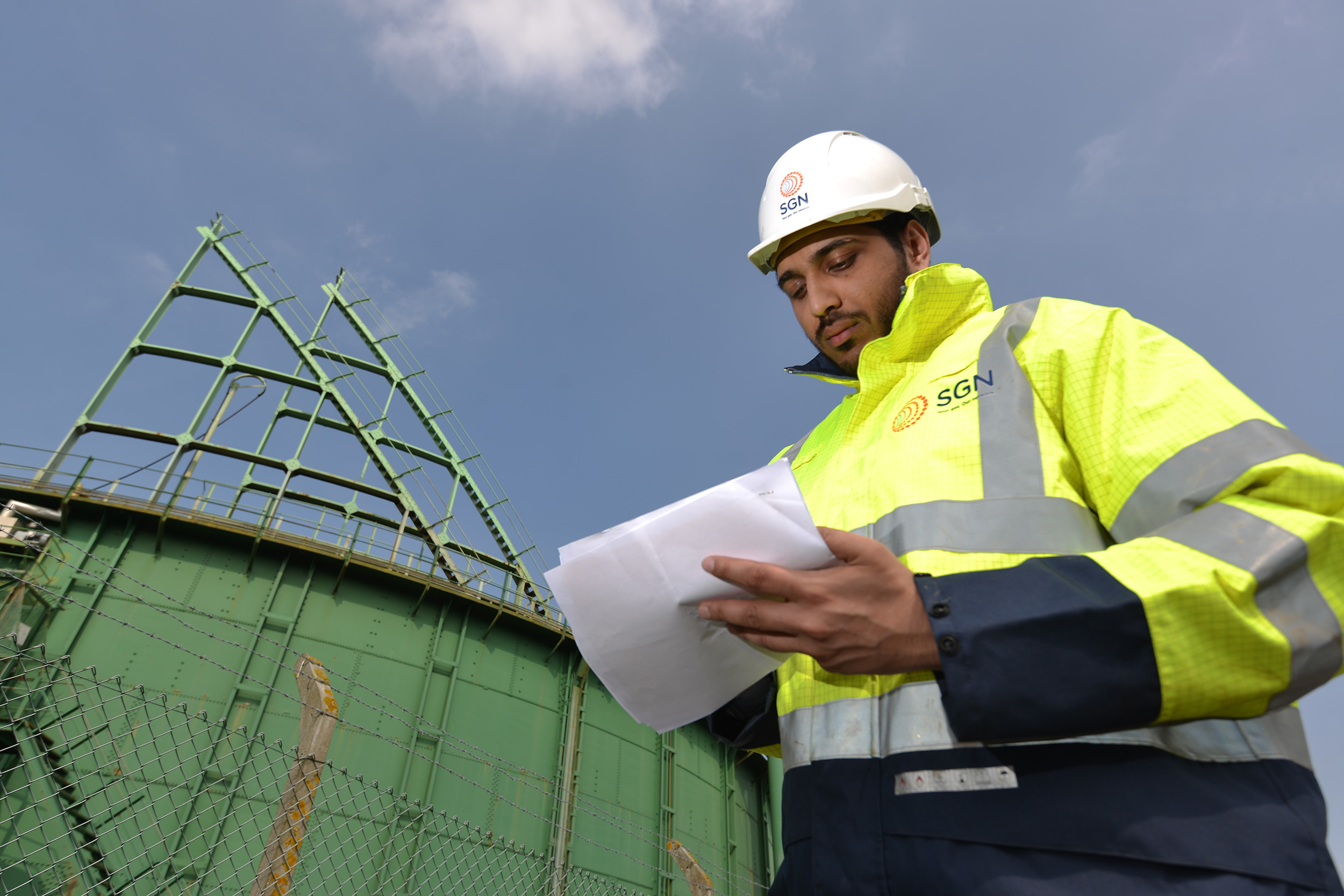 A SGN engineer wearing a high-visibility jacket and hardhat and holding a piece of paper, standing in front of a gas holder
